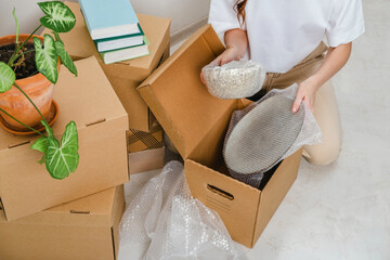 Unrecognized woman wearing white t-shirt sitting on the floor surrounded with carton boxes and packing plates and dishes. Relocating, buying new house. Pack personal stuff from carton boxes. 