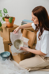 Portrait of woman wearing white t-shirt sitting on the floor surrounded with carton boxes and packing plates and dishes. Relocating, buying new house. Pack personal stuff from carton boxes.
