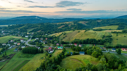 Picturesque countryside landscape in the Bieszczady Mountains captured from above, with green fields, scattered houses, forests, and hills at sunset.