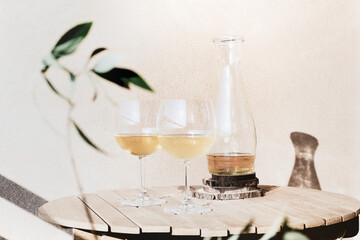 Two glasses of white wine next to an open bottle on a wooden table with an olive branch in the frame on a sunny summer day