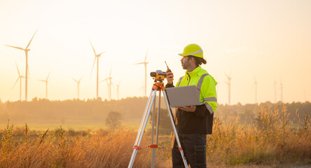 Engineer working on laptop in front of wind turbines for checking wind turbines of the field during beautiful sunset. Alternative energy concept.