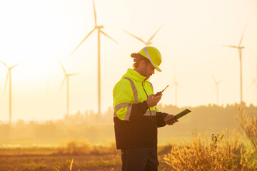 Engineer working on laptop in front of wind turbines for checking wind turbines of the field during beautiful sunset. Alternative energy concept.