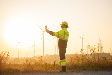 Engineer working on laptop in front of wind turbines for checking wind turbines of the field during beautiful sunset. Alternative energy concept.