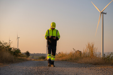 Engineer working on laptop in front of wind turbines for checking wind turbines of the field during beautiful sunset. Alternative energy concept.