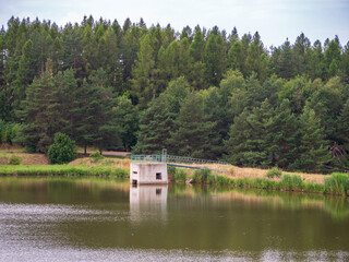 Fototapeta premium Overflow shaft at a fishing pond in Vrbov village near Kežmarok, Slovakia. Calm water, forested surroundings, and peaceful nature scenery.