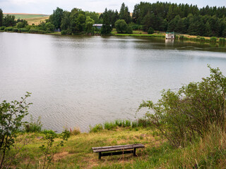 Obraz premium Wooden bench by a quiet pond with forest, fields and a small concrete water structure. Rural scenery near Vrbov, Slovakia, ideal for fishing and nature walks.