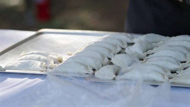 Close-up shot of a cook&rsquo;s hands methodically arranging uncooked empanadas in neat rows on a large tray. Focus on preparation process.