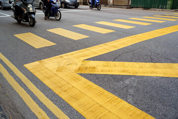 Close-up view of bold yellow pedestrian crosswalk and directional road markings on an urban street with motorcycles and parked vehicles in the background.