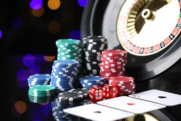 Casino chips, playing cards, roulette wheel and dice on mirror surface against blurred lights,...