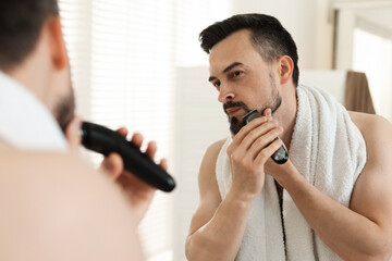 Handsome man shaving with trimmer near mirror at home