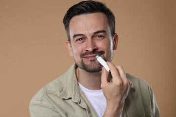 Handsome man applying lip balm onto his lips on light brown background