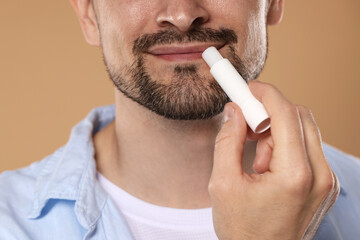 Man applying lip balm onto his lips on light brown background, closeup