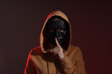 Anonymity. Woman wearing mask and showing hush gesture on dark background