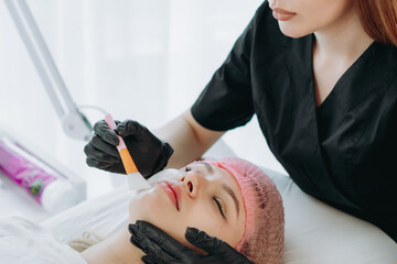professional aesthetician applies a creamy face mask on young woman face using a brush