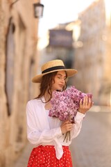 Beautiful woman in hat with bouquet of lilac flowers near building outdoors