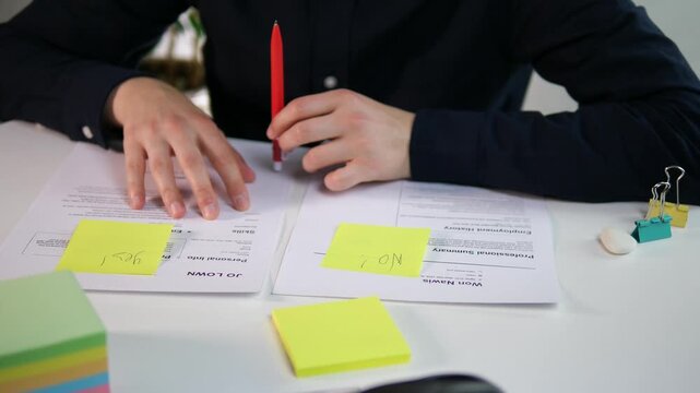 Close-up of a male executive's hands in a dark shirt tapping a pen on a white desk while reviewing resumes. The marked 'Yes' and 'No' notes reveal a critical hiring decision in progress. 