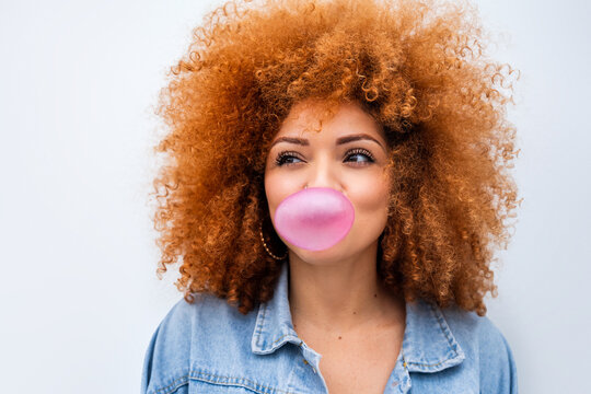 Isolated closeup portrait of smiling ginger teenager girl blowing pink chewing gum. Playful latin young woman feeling positive emotion showing satisfied expression, having fun with bubblegum at wall
