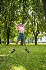 funny happy smiling little girl in glasses in pink top and jeans  jumping in park on sunny summer...