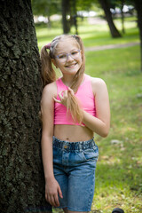 funny happy smiling little girl in glasses in pink top and jeans  posing in park on sunny summer day