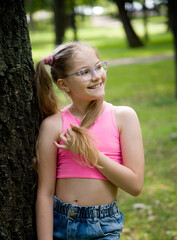funny happy smiling little girl in glasses in pink top and jeans  posing in park on sunny summer day