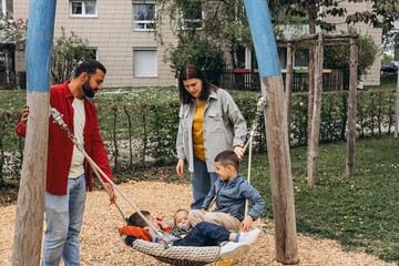 Happy family with two sons playing together on a large rope swing at playground. Parents smiling,...