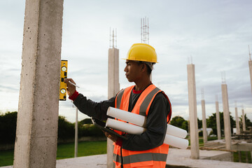 A construction worker wearing a yellow helmet and safety vest examines blueprints on a building...