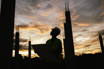 Silhouette of a construction worker holding blueprints and using a phone at a building site during...
