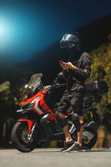 Cool biker checking social media on phone by motorcycle at night, modern lifestyle and confidence, Stylish rider browsing network on device near bike in evening, adventure and freedom