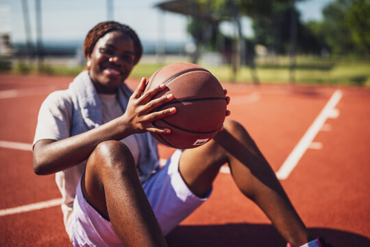 Young black woman is sitting on basketball court. She is holding the ball. Selective focus. - Powered by Adobe