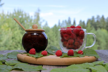 A wooden plate with a glass jar of fresh raspberry jam and a glass mug of ripe raspberries is placed among raspberry leaves on an old wooden table  against a summer forest landscape. 