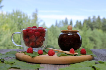 A wooden plate with a glass jar of fresh raspberry jam and a glass mug of ripe raspberries is placed among raspberry leaves on an old wooden table  against a summer forest landscape. 