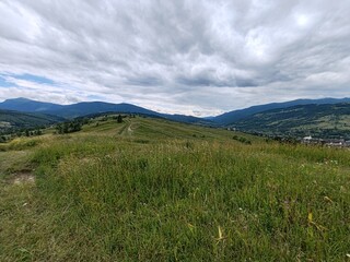 A picturesque summer view of the Carpathian Mountains in Zakarpattia Oblast, Ukraine, featuring rolling green hills, traditional wooden houses, and a vast blue sky with fluffy clouds.