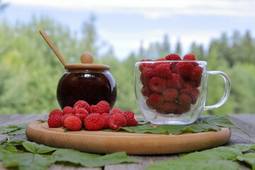 A wooden plate with a glass mug of ripe raspberries, heap of berries and a glass jar of fresh raspberry jam is placed among raspberry leaves on an old wooden table  against a summer forest landscape. 