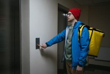 Delivery man wearing a thermal backpack pushing the elevator button while delivering food in an...
