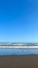A peaceful beach scene with gentle waves meeting the sandy shore beneath a clear blue sky. The calm ocean and expansive sky create a serene coastal atmosphere.