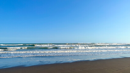 A peaceful beach scene with gentle waves meeting the sandy shore beneath a clear blue sky. The calm ocean and expansive sky create a serene coastal atmosphere.