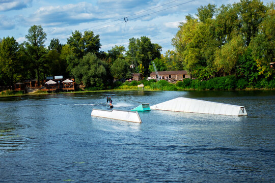 Dynamic shot of a person wakeboarding on a lake with ramps and a cable system, capturing the action, water splash, and surrounding green nature under a cloudy sky. Perfect for active lifestyle