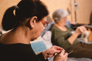 Two senior women knitting indoors, a close-up on the hands of the foreground woman focused on her craft. A cozy scene of domestic hobby and handmade creativity.