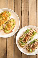 Two plates of delicious rye bread toast topped with avocado, salmon, pistachios, hummus, minced meat, and arugula, arranged on a rustic wooden table