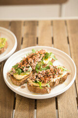 Two slices of whole wheat bread with hummus, arugula and seasoned ground meat are lying on a white plate on a wooden table, with another plate with a toast in the background out of focus