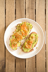 Toasted rye bread slices topped with mashed avocado, cream cheese, smoked salmon, chopped pistachios, and red pepper flakes, served on a white plate on a wooden table
