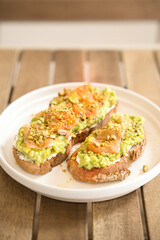 Two slices of toasted whole wheat bread topped with mashed avocado, cream cheese, smoked salmon, crushed pistachios, and spices, served on a white plate