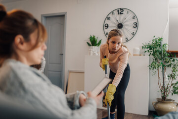 Young girl mopping floor while mother relaxes on sofa using tablet