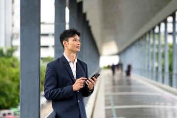 Confident businessman standing on a skywalk, holding smartphone and looking with determination.