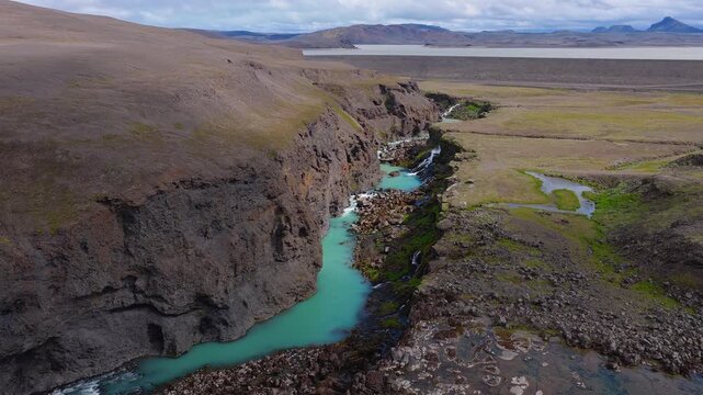 Aerial view of Sigoldugljufur canyon in Iceland with a turquoise river, small waterfall, volcanic terrain, green moss, lake, and distant mountains.