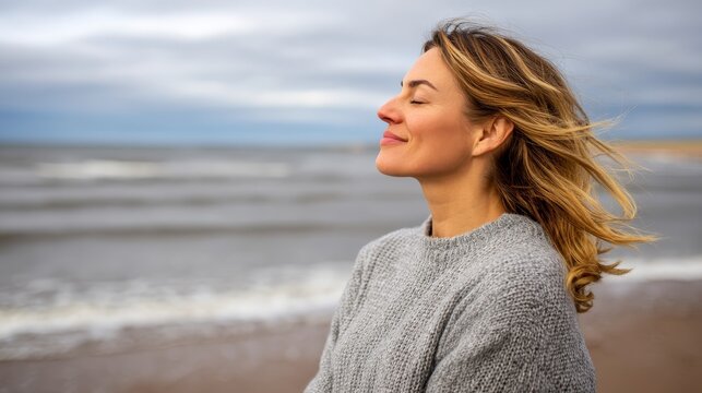 Medium side view of a peaceful woman standing at a breezy coastline with windswept hair, closed eyes, and gentle waves crashing under cloudy skies, creating a serene and tranquil atmosphere