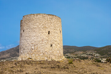 Naklejka premium A historic stone tower stands on a hillside under a clear blue sky. The structure is cylindrical with a rough texture, surrounded by rocky terrain and distant mountains.