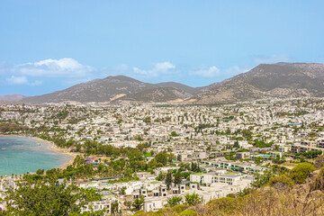 A scenic view of a coastal town with a sandy beach, clear blue water, and distant mountains under a...