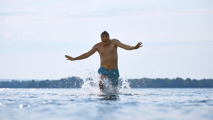 Older man running into cold water with surprised facial expression. Concept of midlife spontaneity,...