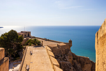 Obraz premium The view from Castillo de Santa Barbara in Alicante showcases a stunning coastline, with vibrant blue waters and palm trees surrounding historical architecture under a clear sky.